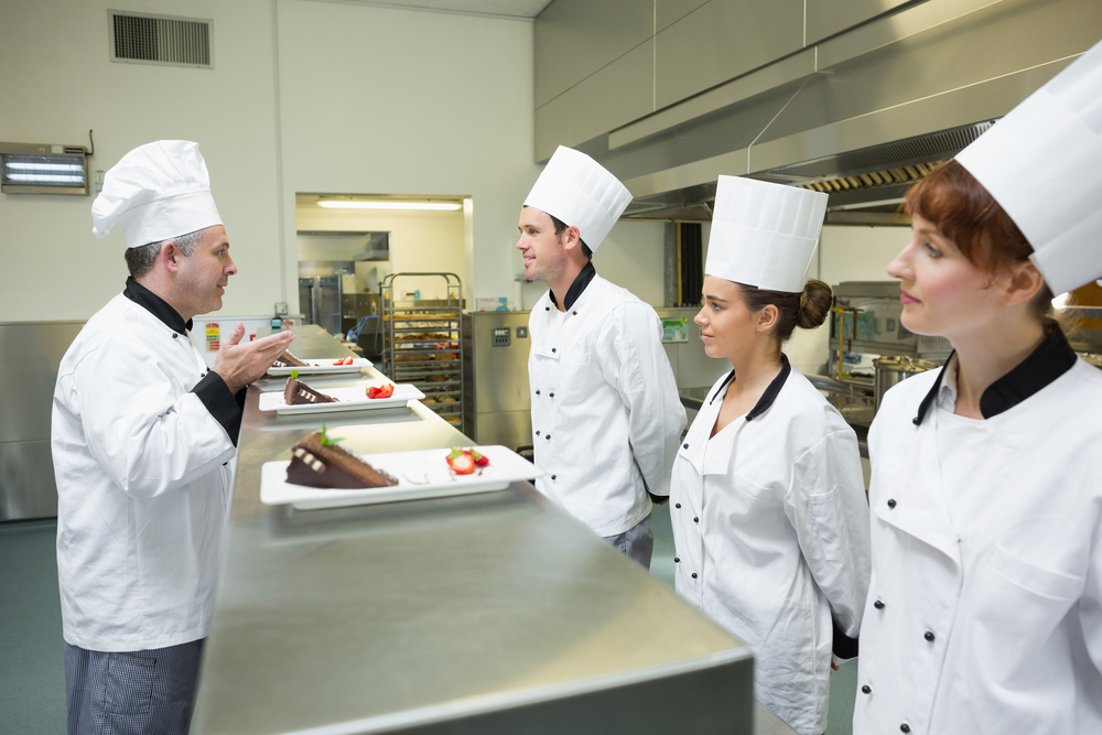 Three chefs presenting their dessert plates to the head chef in busy kitchen